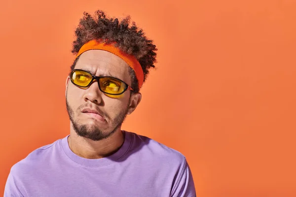 Homem americano africano pensativo em óculos de sol e headband olhando para longe no fundo laranja, grimace — Fotografia de Stock