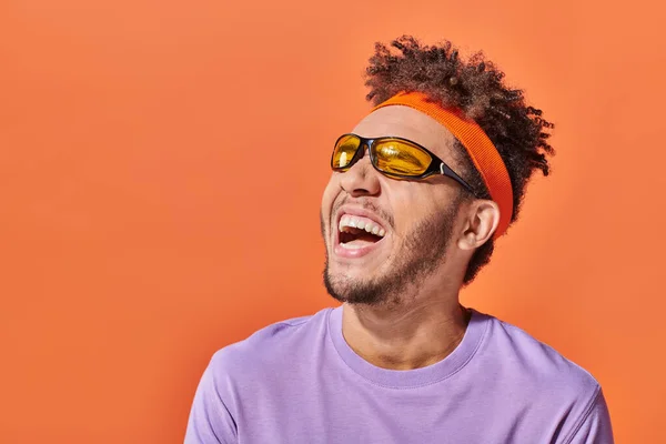 Africano americano uomo in occhiali da sole e fascia ridere e guardando lontano su sfondo arancione — Foto stock