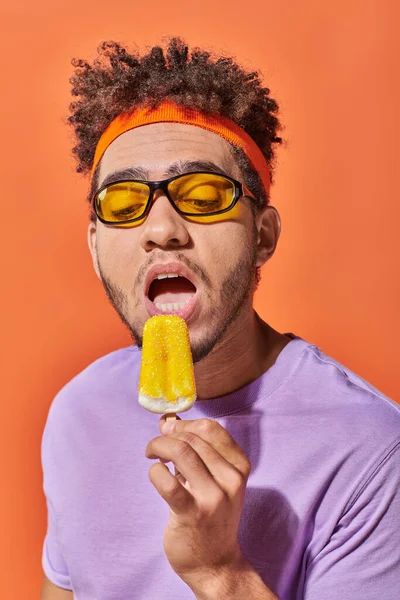 Hombre afroamericano en gafas de sol y diadema comiendo helado afrutado en verano sobre fondo naranja - foto de stock