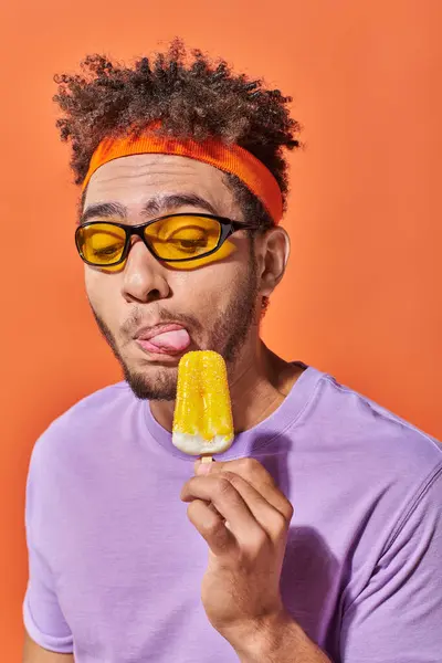 Hombre afroamericano en gafas de sol y diadema lamiendo helado afrutado sobre fondo naranja - foto de stock