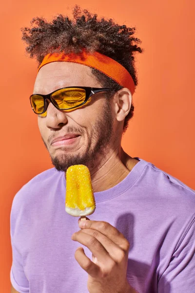 Homem americano africano em óculos de sol e fita de cabeça sorrindo de sorvete azedo em pano de fundo laranja — Fotografia de Stock