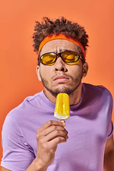 Afrikanisch-amerikanischer Mann mit Sonnenbrille und Stirnband, der nach fruchtigem Eis auf orangefarbenem Hintergrund riecht — Stockfoto