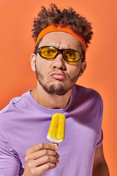 Hombre afroamericano emocional en gafas de sol y diadema celebración de helado afrutado en naranja - foto de stock