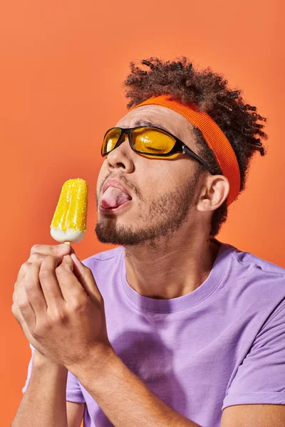 Uomo afroamericano in occhiali da sole e fascia leccare gelato fruttato su sfondo arancione, lingua — Foto stock
