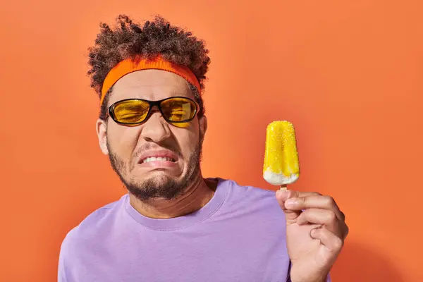 Displeased african american man in sunglasses and headband looking at frozen ice cream on orange — Stock Photo