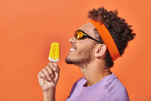 Hombre afroamericano feliz en gafas de sol y diadema con helado congelado sobre fondo naranja - foto de stock