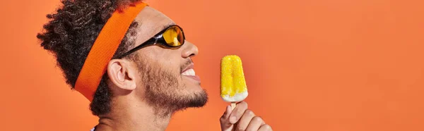Heureux homme afro-américain en lunettes de soleil tenant crème glacée congelée sur fond orange, bannière — Photo de stock