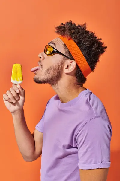 Hombre afroamericano en gafas de sol y diadema lamiendo helado congelado sobre fondo naranja, lengua - foto de stock
