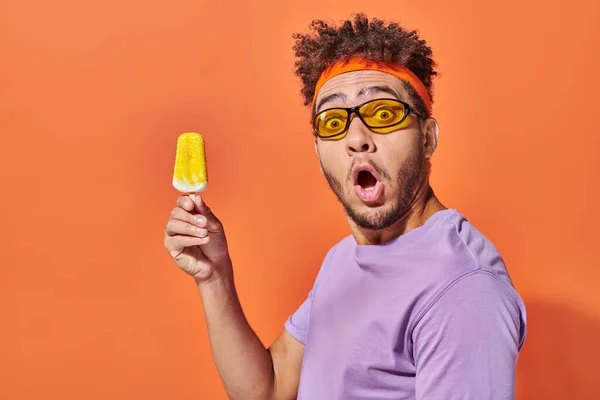 Chocado afro-americano homem em óculos de sol e headband segurando congelado sorvete no fundo laranja — Fotografia de Stock