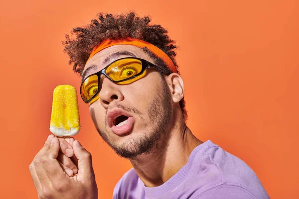 Chocado homem americano africano em óculos de sol segurando sorvete congelado no fundo laranja — Fotografia de Stock