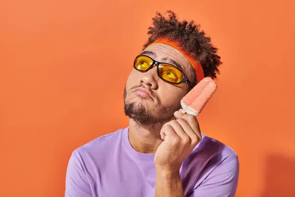 Hombre afroamericano pensativo en gafas de sol celebración de helado con aspersiones sobre fondo naranja - foto de stock