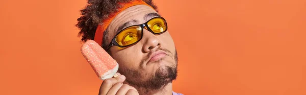 Hombre afroamericano pensativo en gafas de sol celebración de helado con aspersiones en naranja, pancarta - foto de stock