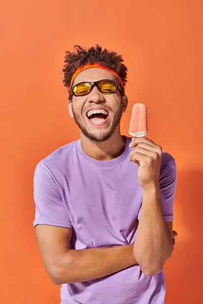 Hombre afroamericano feliz en gafas de sol sosteniendo helado con aspersiones sobre fondo naranja - foto de stock