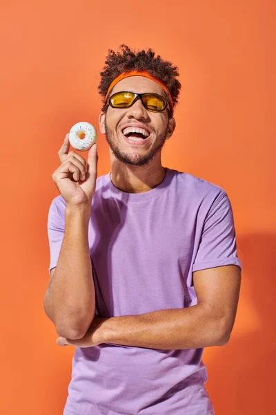 Happy young african american man in sunglasses holding small tasty donut on orange background — Stock Photo