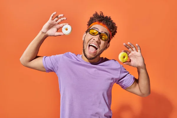 Щасливий молодий афроамериканський чоловік в сонцезахисних окулярах тримає маленькі смачні пончики на помаранчевому фоні — стокове фото