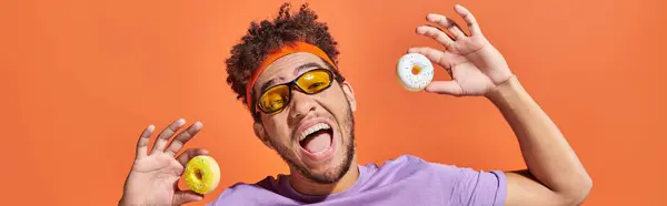 Feliz jovem afro-americano em óculos de sol segurando pequenos donuts no fundo laranja, banner — Fotografia de Stock
