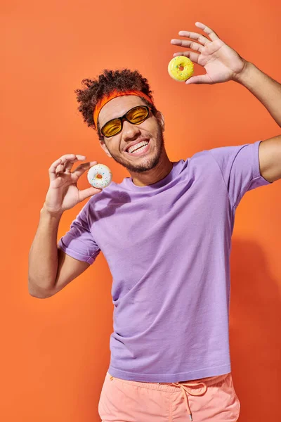 Щасливий молодий афроамериканський чоловік в сонцезахисних окулярах тримає пончики розміром з укус на помаранчевому фоні — стокове фото