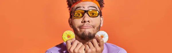 Funny african american man in sunglasses holding bite-sized donuts on orange background, banner — Stock Photo