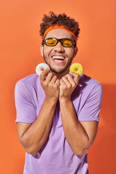 Joyeux jeune homme afro-américain dans des lunettes de soleil tenant des beignets de taille morsure sur fond orange — Photo de stock