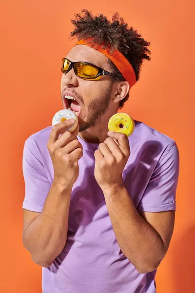 Joven afroamericano hombre en gafas de sol mordiendo sabrosa rosquilla azucarada sobre fondo naranja - foto de stock