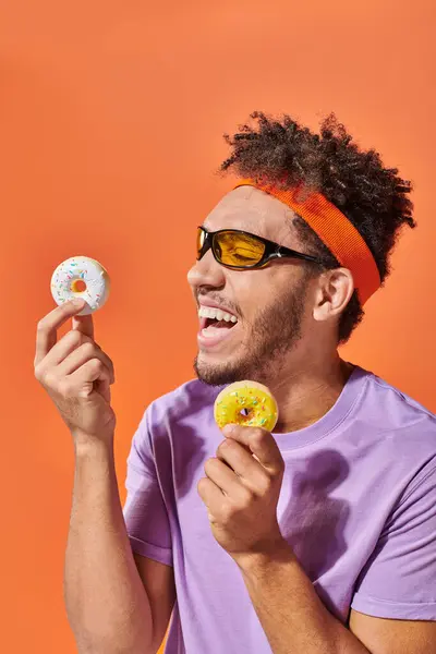Happy african american man in sunglasses holding bite-sized donuts and laughing on orange background — Stock Photo