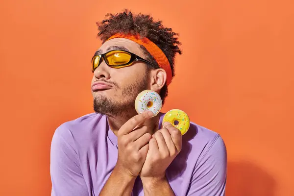 African american man in sunglasses holding bite-sized donuts on orange background, grimace — Stock Photo