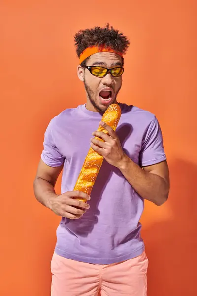 Joven afroamericano hombre en gafas de sol y diadema mordiendo baguette fresca sobre fondo naranja - foto de stock