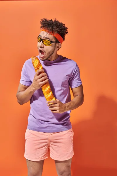 Смешной африканский американец в солнцезащитных очках и ободок, кусающий свежий багет на оранжевом фоне — стоковое фото