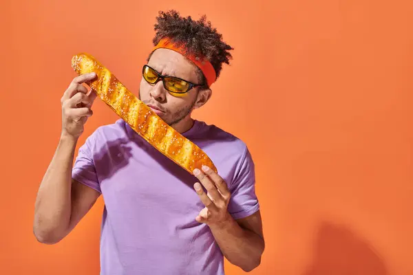Young african american man in sunglasses and headband smelling fresh baguette on orange background — Stock Photo