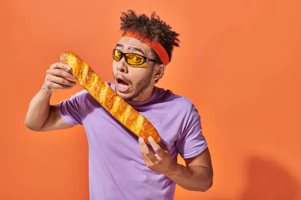 Uomo africano americano in occhiali da sole e fascia pungente baguette fresca su sfondo arancione, panetteria — Foto stock