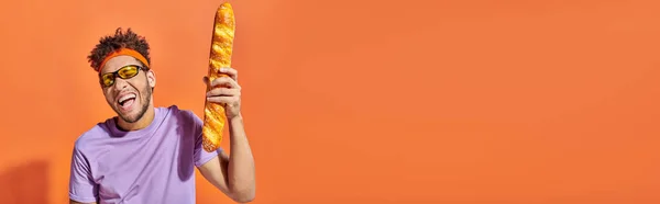 Hombre afroamericano feliz en gafas de sol sosteniendo baguette fresca sobre fondo naranja, bandera - foto de stock
