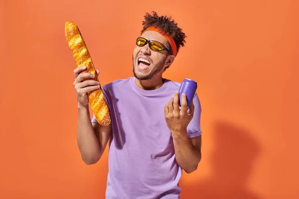 Feliz homem americano africano em óculos de sol segurando baguete fresco e refrigerante no fundo laranja — Fotografia de Stock