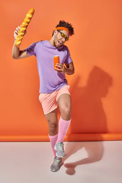 Hombre afroamericano excitado en gafas de sol sosteniendo baguette fresca y soda sobre fondo naranja - foto de stock