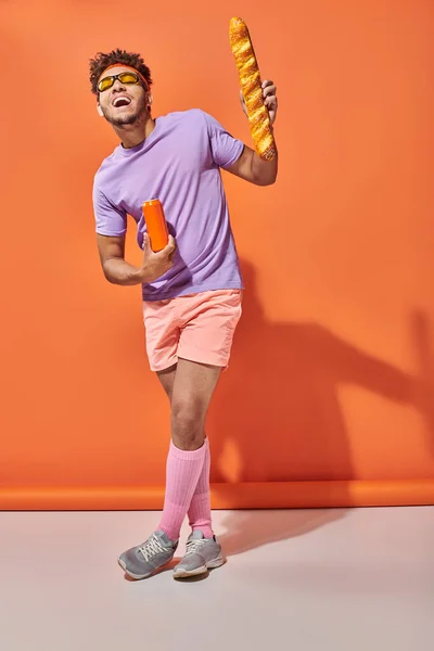 Веселий афроамериканський чоловік в сонцезахисних окулярах тримає свіжий багет і соду на помаранчевому фоні — стокове фото