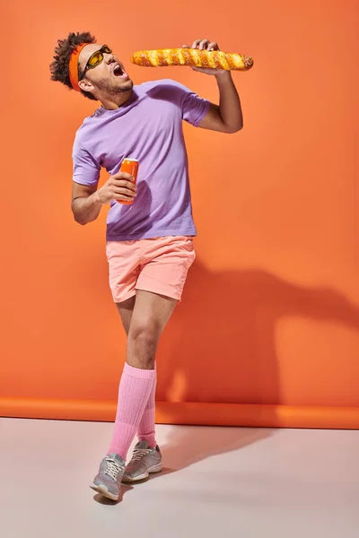 Hombre afroamericano en gafas de sol comiendo baguette fresca y sosteniendo soda sobre fondo naranja - foto de stock