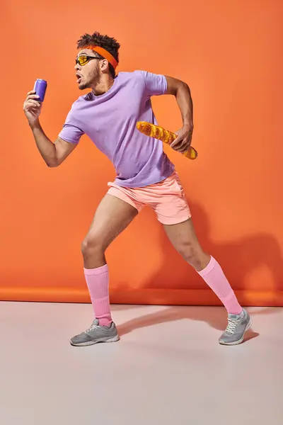 Jeune homme afro-américain en lunettes de soleil tenant baguette fraîche et soda sur fond orange — Photo de stock