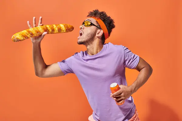 Hombre afroamericano apasionado en gafas de sol comiendo baguette fresca y sosteniendo soda en naranja - foto de stock