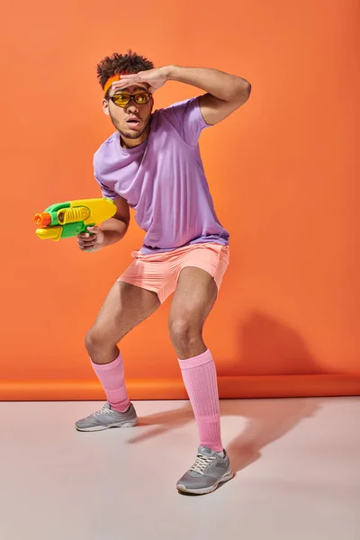 Young african american guy in sunglasses looking away while holding water gun on orange background — Stock Photo