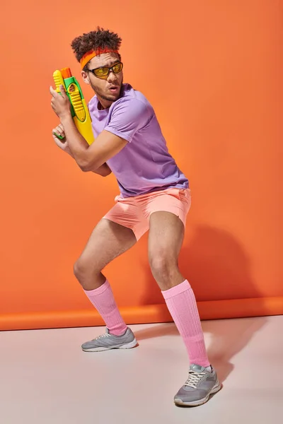 Tensed african american guy in sunglasses looking away while holding water gun on orange background — Stock Photo