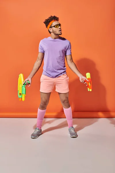 Tensed african american man in sunglasses looking away while holding water guns on orange background — Stock Photo