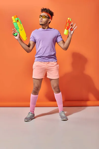 Tensed african american guy in sunglasses looking away while holding water guns on orange background — Stock Photo