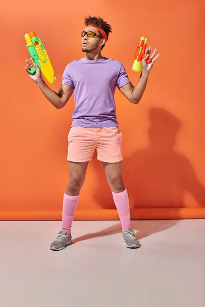 Tipo afroamericano tenso en gafas de sol mirando hacia otro lado mientras sostiene las armas de agua sobre fondo naranja - foto de stock