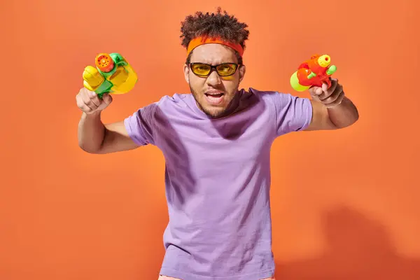 Angespannte afrikanisch-amerikanische Fella in Sonnenbrille mit Wasserpistolen auf orangefarbenem Hintergrund, Sommerspaß — Stockfoto