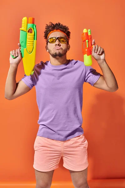 Напружений афроамериканський чоловік в сонцезахисних окулярах тримає водяні гармати на помаранчевому фоні, літні забави — стокове фото