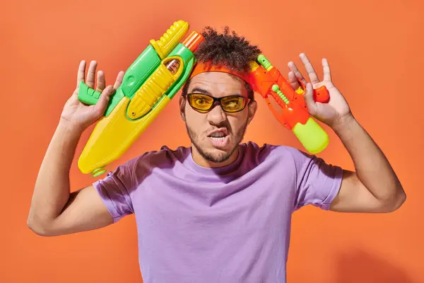 Tensed african american man in sunglasses  holding water guns on orange background, grimace — Stock Photo