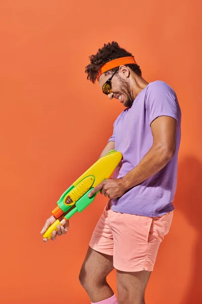 Hombre afroamericano tenso en gafas de sol sosteniendo pistola de agua sobre fondo naranja, mueca - foto de stock
