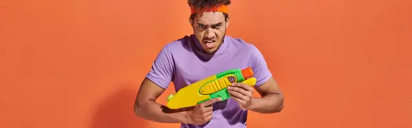 African american man in headband playing water fight with toy gun on orange background, banner — Stock Photo