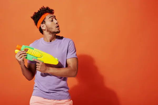 Curly african american man in headband playing water fight with toy gun on orange background — Stock Photo