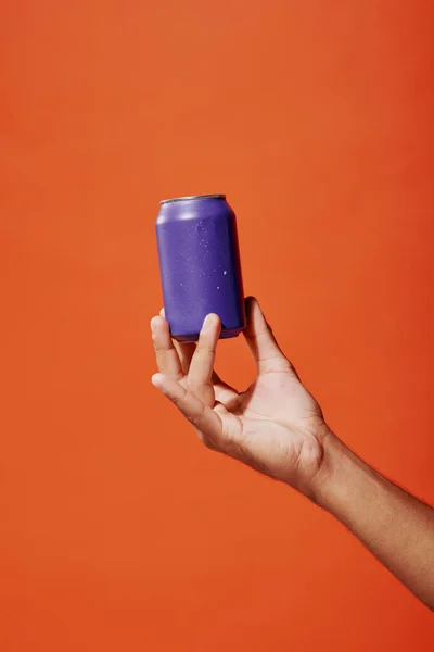 Tiro recortado de la persona que sostiene lata de refresco púrpura en la mano sobre fondo naranja, bebida carbonatada - foto de stock