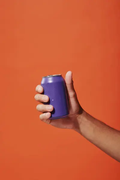 Foto recortada de la persona que sostiene lata de refresco púrpura en la mano sobre fondo naranja, bebida carbonatada - foto de stock